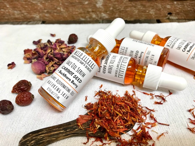 May include: A close-up of a small glass bottle with a dropper filled with a golden liquid. The bottle is labeled "Wild Elixirs Face Oil Serum Carrot Seed + Buckthorn Berry". The bottle is surrounded by dried rose petals and saffron.