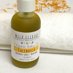 Puede incluir: Una botella de vidrio transparente con una etiqueta blanca que dice "WILD ELIXIR PURE NATURALLY NOURISHING SKINCARE Organic Baby Oil CALENDULA UNSCENTED/ GENTLE/ NOURISHING + VITAMIN E NT.WT. 2 fl oz/ 59 ml". La botella est&aacute; llena de un l&iacute;quido amarillo y est&aacute; sobre una toalla blanca.
