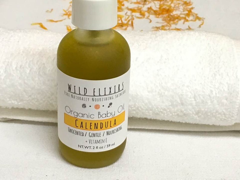 Puede incluir: Una botella de vidrio transparente con una etiqueta blanca que dice "WILD ELIXIR PURE NATURALLY NOURISHING SKINCARE Organic Baby Oil CALENDULA UNSCENTED/ GENTLE/ NOURISHING + VITAMIN E NT.WT. 2 fl oz/ 59 ml". La botella est&aacute; llena de un l&iacute;quido amarillo y est&aacute; sobre una toalla blanca.