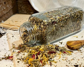 May include: A glass jar filled with a blend of dried herbs and flowers, including yellow calendula petals, green herbs, and pink rose petals. The jar is tipped over, spilling the contents onto a white surface.