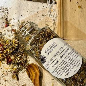 May include: A glass jar filled with dried herbs and flowers, labeled "Wild Elixirs Raw Skincare Essentials Soothe Herbal Bath Soak Orange + Ylang-Ylang" with a list of ingredients including calendula, yarrow, chickweed, pink Himalayan sea salt, lavender, St. John's Wort, comfrey leaf, rose petals, chamomile, hibiscus, lavender, jojoba oil, non-GMO natural vitamin E, essential oils of sweet orange and ylang-ylang, and wild crafted. The jar is sitting on a white fabric surface with a wooden spoon next to it.