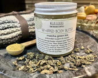 May include: A white jar with a green lid labeled "Wild Elixirs Raw Skincare Essentials Whipped Body Butter Matcha Green Tea" with a list of ingredients including mango butter, shea butter, coconut oil, green tea, argan oil, hemp seed oil, and sweet orange. The jar is sitting on a gray surface with green tea leaves scattered around it.