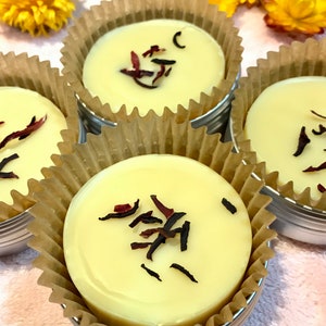 Peut inclure: Quatre petits contenants ronds de beurre corporel blanc avec des pétales de fleurs rouges séchées sur le dessus. Les contenants sont en métal et ont une doublure en papier brun.