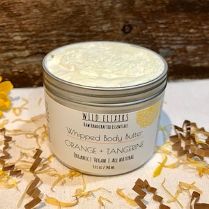May include: A silver tin of whipped body butter with a white label that reads "WILD ELIXIRS RAW HANDCRAFTED ESSENTIALS Whipped Body Butter ORANGE + TANGERINE ORGANICI VEGAN | ALL NATURAL 5 fl oz / 148 ml"