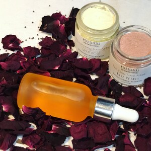 May include: Three skincare products with rose petals: a clear glass bottle with a dropper filled with orange liquid, a white jar with a white lid labeled "Rejuvenating Face Whip Wild Rose", and a pink jar with a black lid labeled "Pink Clay Face Mask Rose Petals".