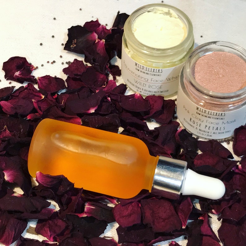 May include: Three skincare products with rose petals: a clear glass bottle with a dropper filled with orange liquid, a white jar with a white lid labeled "Rejuvenating Face Whip Wild Rose", and a pink jar with a black lid labeled "Pink Clay Face Mask Rose Petals".