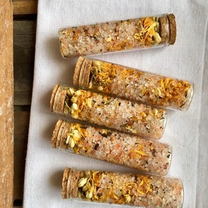 May include: Five glass tubes filled with bath salts and dried flowers, each sealed with a cork. The salts are a mix of pink and white crystals, with yellow and white flower petals. The tubes rest on a white cloth.