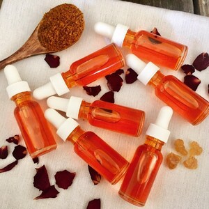 May include: Seven small amber glass bottles with droppers filled with a bright orange liquid. The bottles are arranged on a white surface with rose petals and a small pile of brown sugar.