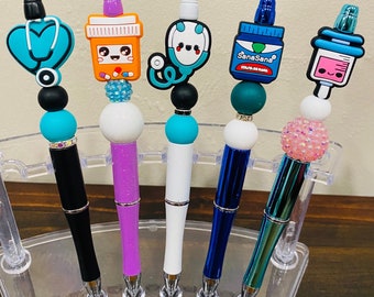 May include: Five colorful pens with different medical-themed toppers, including a stethoscope, a pill bottle, a syringe, and a bottle of hand sanitizer. The pens are displayed in a clear plastic stand.