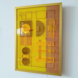 May include: Abstract art piece in a gold frame. The artwork features geometric shapes in yellow, brown, and clear acrylic, with a wooden panel on the right side. The design includes circles, squares, and lines, creating a modern aesthetic.