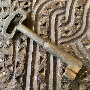 May include: A weathered metal key with a rectangular handle and a ridged shaft. The key is a dull silver colour and rests on a carved wooden surface with geometric and curved patterns. The key's head has a distinctive, open design.