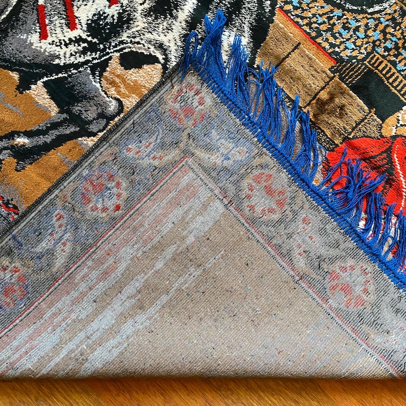 K&ouml;nnte beinhalten: Ein Abschnitt eines dekorativen Teppichs mit detailliertem Design. Der Teppich hat eine Farbpalette aus Beige, Braun, Schwarz und Rot mit blauen Fransen. Das Design umfasst einen Ritter auf einem Pferd und geometrische Muster.