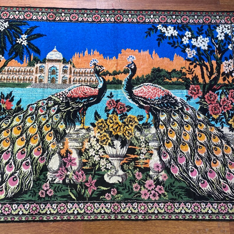 Peut inclure: Tapisserie d&eacute;corative repr&eacute;sentant deux paons aux plumes vibrantes, debout sur un mur surplombant un plan d'eau. L'arri&egrave;re-plan comprend un palais, des arbres et des fleurs, avec un ciel bleu et une bordure florale.