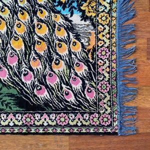 Peut inclure: Une section d'un tapis color&eacute; sur le th&egrave;me du paon. Le tapis pr&eacute;sente un fond noir avec des motifs de plumes de paon roses, oranges et jaunes. La bordure pr&eacute;sente un motif floral avec des accents roses et verts, et le bord est orn&eacute; de franges bleues.