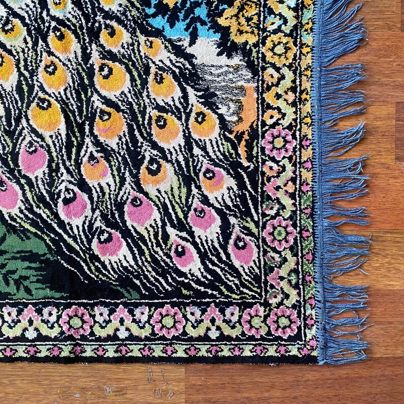 Peut inclure: Une section d'un tapis color&eacute; sur le th&egrave;me du paon. Le tapis pr&eacute;sente un fond noir avec des motifs de plumes de paon roses, oranges et jaunes. La bordure pr&eacute;sente un motif floral avec des accents roses et verts, et le bord est orn&eacute; de franges bleues.