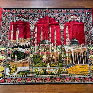 May include: A rectangular prayer rug with a red central field depicting detailed illustrations of Islamic architecture. The rug has a black border with floral patterns and red fringe. It is placed on a wooden surface.