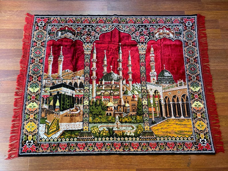 May include: A rectangular prayer rug with a red central field depicting detailed illustrations of Islamic architecture. The rug has a black border with floral patterns and red fringe. It is placed on a wooden surface.