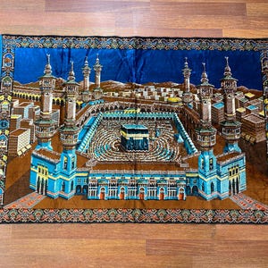 May include: A rectangular prayer rug with a detailed depiction of a religious site. The rug features a central structure surrounded by minarets and buildings, with a blue border and fringe.