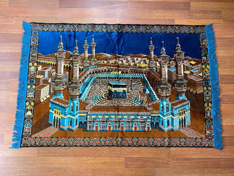 May include: A rectangular prayer rug with a detailed depiction of a religious site. The rug features a central structure surrounded by minarets and buildings, with a blue border and fringe.