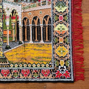 May include: A rectangular rug with a detailed architectural design in various colours. The rug features a yellow and brown courtyard, arched columns, and a red fringe border. The border has floral and geometric patterns in red, green, and yellow.