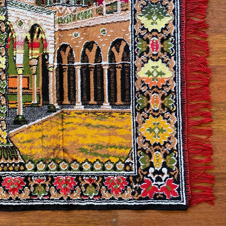 May include: A rectangular rug with a detailed architectural design in various colours. The rug features a yellow and brown courtyard, arched columns, and a red fringe border. The border has floral and geometric patterns in red, green, and yellow.