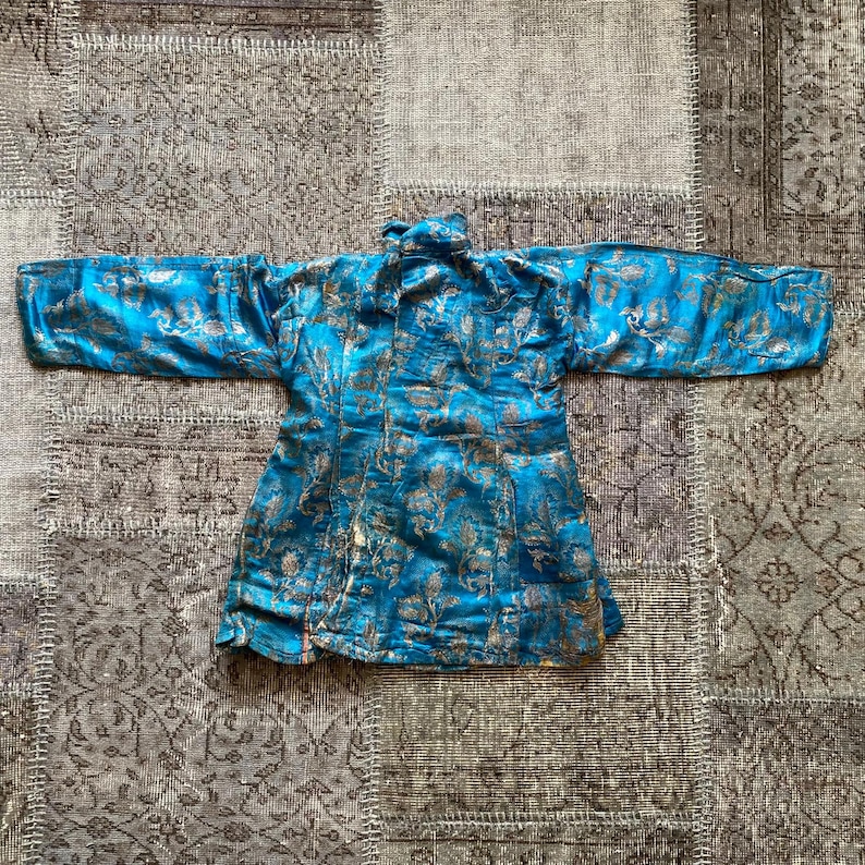 Puede incluir: Una chaqueta de brocado azul y dorado con cuello mandar&iacute;n y mangas largas. La chaqueta est&aacute; decorada con un patr&oacute;n floral.