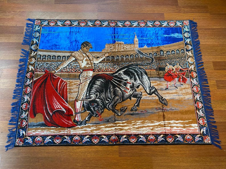 K&ouml;nnte beinhalten: Gewebte Tapisserie mit einer Stierszenen-Darstellung. Ein Matador in traditioneller Kleidung steht einem schwarzen Stier in einer Arena gegen&uuml;ber. Die Szene umfasst ein rotes Kap, weitere Matadore und einen detaillierten Hintergrund der Arena und des Himmels. Der Rand ist blau mit floralen Mustern.