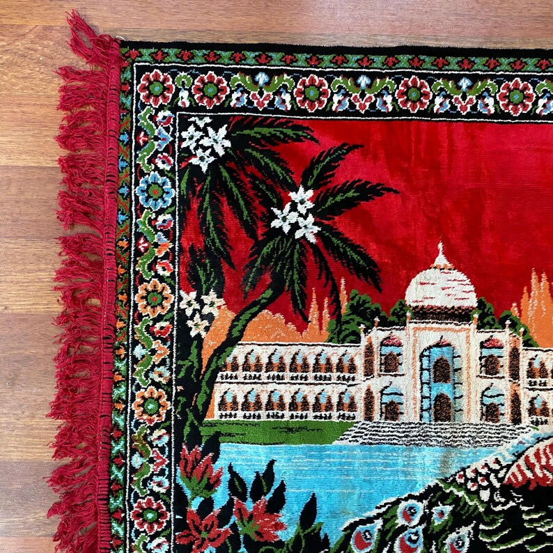 May include: A red decorative rug featuring a detailed illustration of a palace, palm trees, and a peacock. The rug has a black border with floral patterns and red fringe. The rug is made of a soft material.