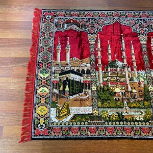 May include: A red prayer mat featuring a detailed depiction of a mosque and the Kaaba. The mat has a central design with minarets and domes, surrounded by floral borders in green, yellow, and black. The edges have a red fringe.