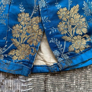 May include: Close-up of the bottom edge of a blue fabric with a gold floral pattern. The fabric has a seam down the center and is folded over at the bottom edge.