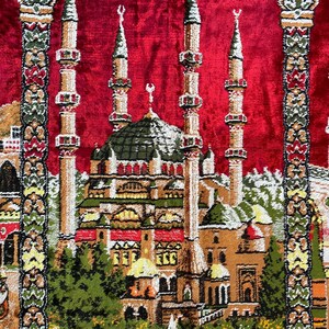 May include: A red prayer rug with a detailed mosque design. The mosque features multiple minarets and a central dome, rendered in brown, green, and beige. The design is set against a red background, with decorative borders framing the central image.