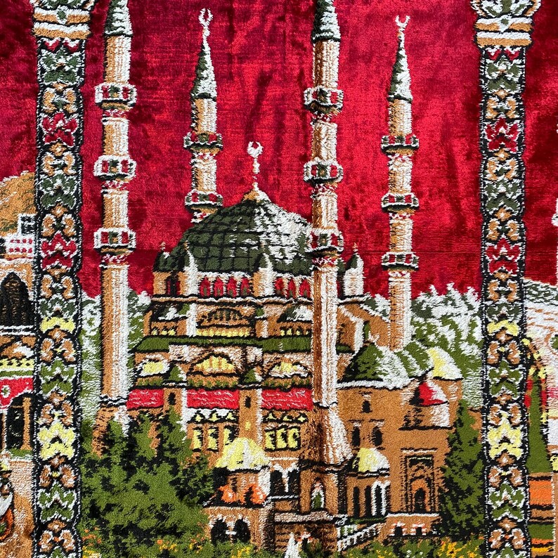 May include: A red prayer rug with a detailed mosque design. The mosque features multiple minarets and a central dome, rendered in brown, green, and beige. The design is set against a red background, with decorative borders framing the central image.