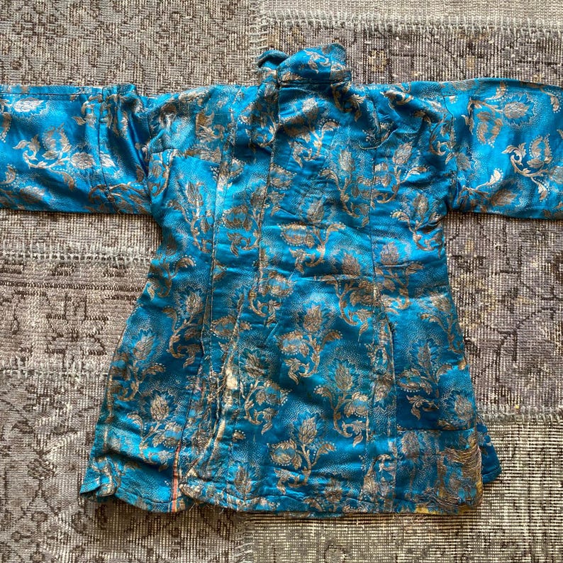 Puede incluir: Una chaqueta azul de brocado con un patr&oacute;n floral y detalles dorados. La chaqueta tiene un cuello mandar&iacute;n y una silueta larga y fluida.