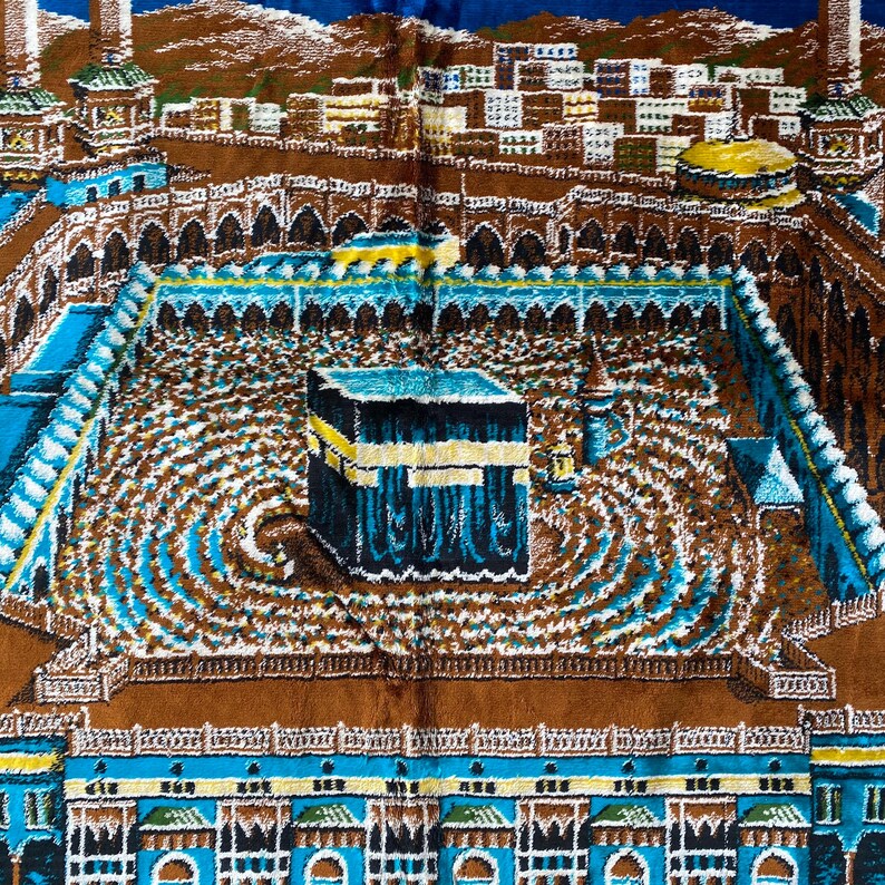 May include: A detailed, colorful prayer rug depicting the Kaaba in Mecca. The rug features a central black cube with gold accents, surrounded by a courtyard with concentric circles. The design includes architectural elements in shades of brown, blue, and white.