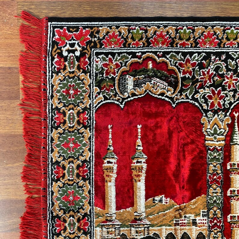 May include: A red prayer rug with a detailed design featuring a mosque and floral patterns. The rug has a black border with red and green floral accents and a red fringe.