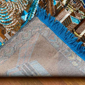 May include: An area rug with a geometric pattern in shades of brown, blue, and beige. The rug has a blue fringe along one edge and a detailed border. The design features repeating shapes and patterns.