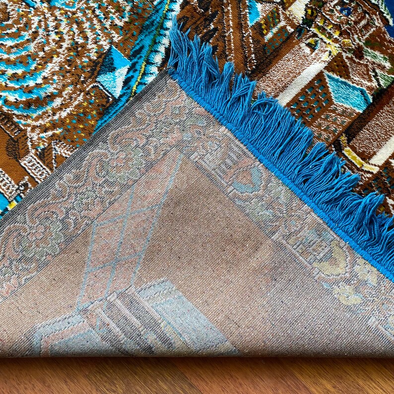 May include: An area rug with a geometric pattern in shades of brown, blue, and beige. The rug has a blue fringe along one edge and a detailed border. The design features repeating shapes and patterns.