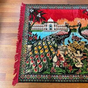 May include: A decorative rug with a peacock design. The rug has a red background with a detailed illustration of a palace, peacocks, and floral arrangements. The rug's border and fringe are red.