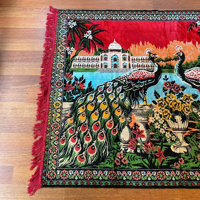 May include: A decorative rug with a peacock design. The rug has a red background with a detailed illustration of a palace, peacocks, and floral arrangements. The rug's border and fringe are red.