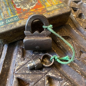 May include: A small, antique metal lock with a matching key, connected by a green string. The lock is dark gray and sits on a carved wooden surface. A vintage cigarette tin is in the background.