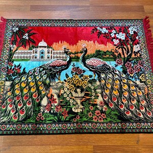 May include: A rectangular rug with a peacock design. Two peacocks face each other, their colourful tail feathers displayed. The rug features a red border with fringe, and a detailed landscape background.