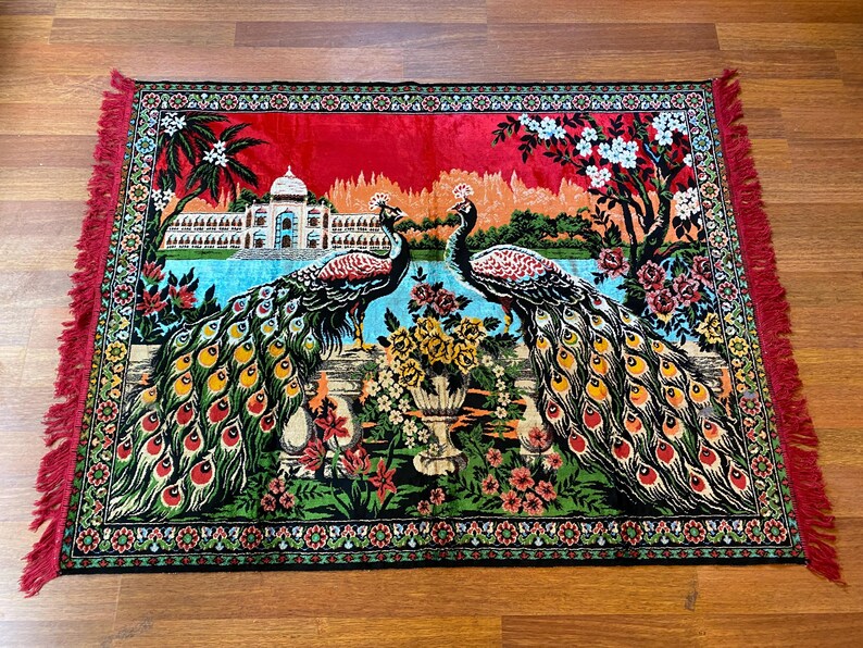 May include: A rectangular rug with a peacock design. Two peacocks face each other, their colourful tail feathers displayed. The rug features a red border with fringe, and a detailed landscape background.