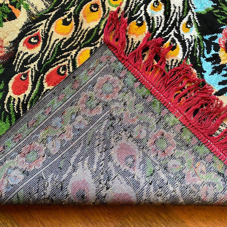May include: A patterned rug with a peacock feather design. The rug has a black base with bright red, yellow, green, and blue details. The underside of the rug is visible, displaying a floral pattern and a red fringe.
