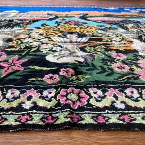 Peut inclure: Gros plan sur un tapis d&eacute;taill&eacute; avec un motif floral et paon. Le tapis pr&eacute;sente un fond noir avec des fleurs roses et jaunes, un feuillage vert et une bordure avec des motifs floraux roses. Le haut est bleu et orange.
