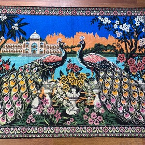 Peut inclure: Tapisserie d&eacute;corative repr&eacute;sentant deux paons aux plumes vibrantes, sur fond de palais, d'eau et de compositions florales. La tapisserie a une frange bleue et une bordure d&eacute;taill&eacute;e &agrave; motifs floraux.