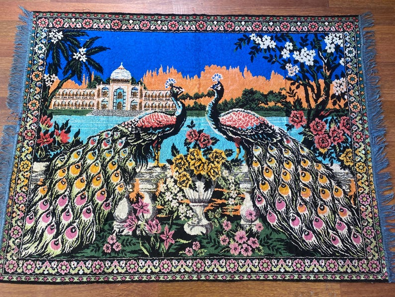 Peut inclure: Tapisserie d&eacute;corative repr&eacute;sentant deux paons aux plumes vibrantes, sur fond de palais, d'eau et de compositions florales. La tapisserie a une frange bleue et une bordure d&eacute;taill&eacute;e &agrave; motifs floraux.