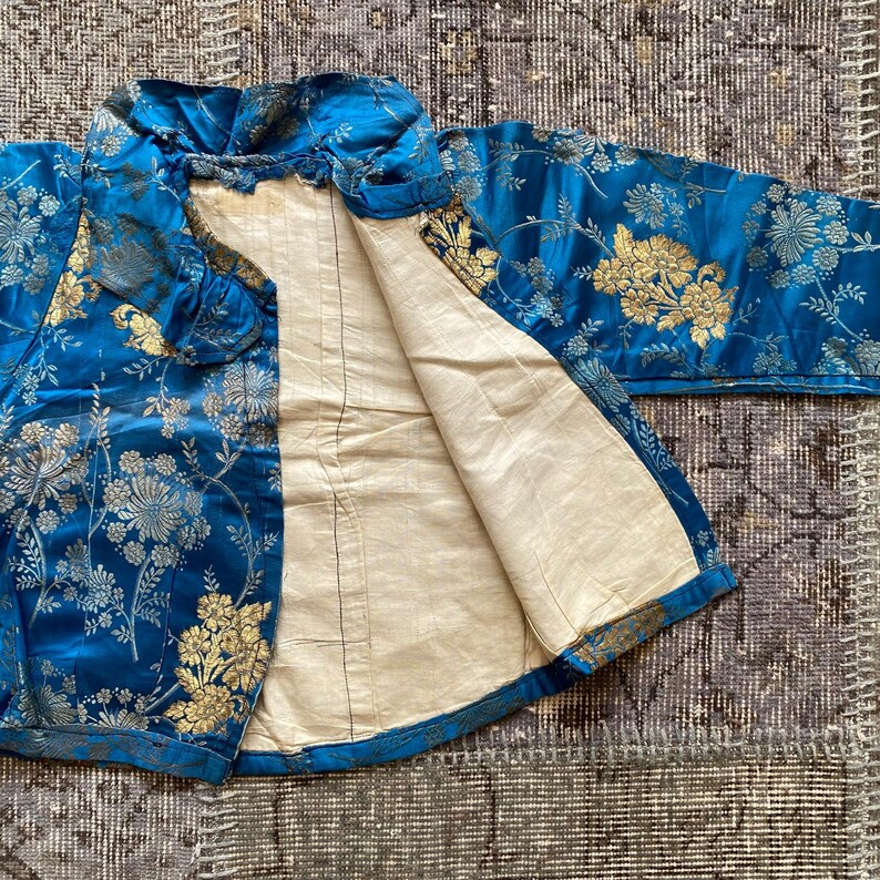 May include: A blue silk jacket with a gold floral pattern. The jacket is lined with a white fabric and has a mandarin collar.