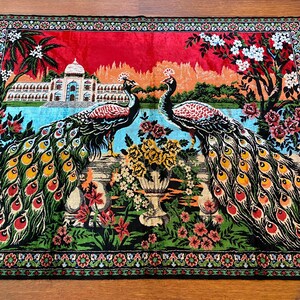 May include: A decorative textile featuring two peacocks with vibrant, fanned tail feathers. The design includes a palace, flowers, and a red sky. The border is detailed with floral patterns, creating a colourful and ornate tapestry.
