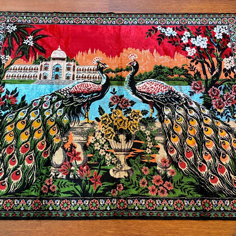 May include: A decorative textile featuring two peacocks with vibrant, fanned tail feathers. The design includes a palace, flowers, and a red sky. The border is detailed with floral patterns, creating a colourful and ornate tapestry.