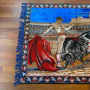 K&ouml;nnte beinhalten: Wandteppich mit einer Stierszenen-Darstellung. Ein Matador in traditioneller Kleidung steht einem angreifenden schwarzen Stier mit rotem Umhang gegen&uuml;ber. Der Hintergrund zeigt ein Stadion und blauen Himmel, mit dekorativem Rand und blauer Fransen.